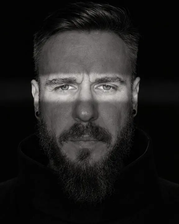 Black-and-white close portrait of a bearded man, a narrow band of hard light crossing his eyes, the rest in black