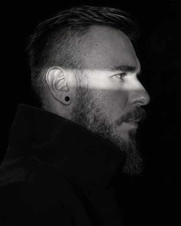 Profile of a man with a strip of light across his eye and cheekbone, a black funnel collar shape below