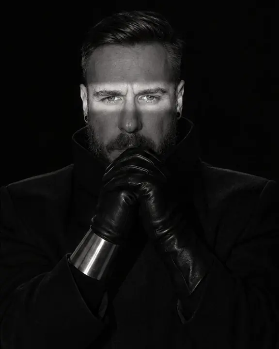 A man raises black-gloved hands near his face inside a band of light, a silver cuff gleaming below his lit eyes