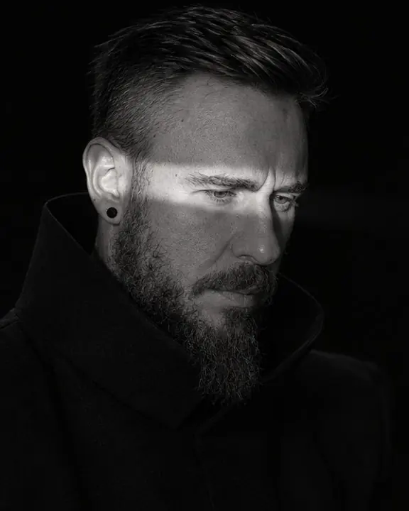 Three-quarter view of a bearded man looking slightly down, a blade of light slicing across his eyes and nose bridge