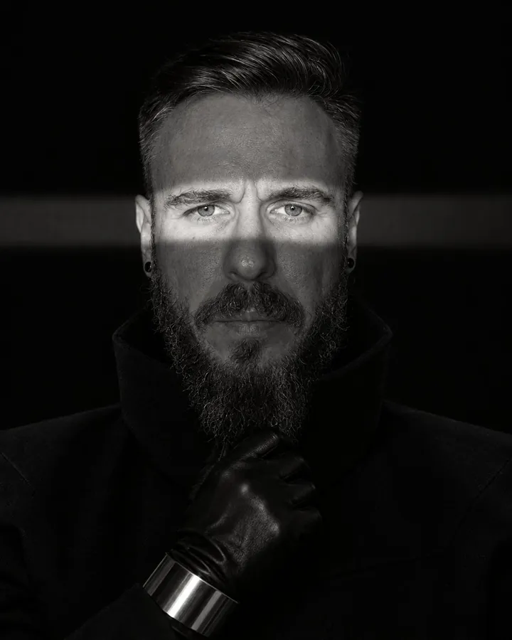 Black-and-white close portrait of a bearded man, a narrow band of hard light crossing his eyes, the rest in black