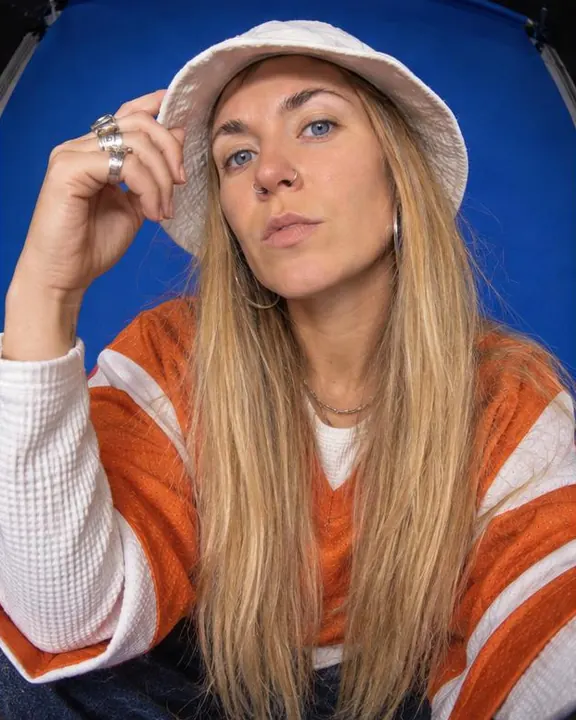 A woman with long blond hair raises a ringed hand beside her face, bucket hat tipped back, a curved paper seam behind