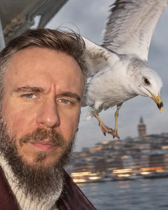 A seagull frozen by flash right beside a bearded man's face, the Istanbul shoreline smeared behind