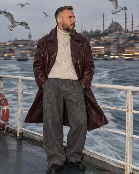 A man in a burgundy leather coat stands at a ferry railing, coat blown by wind, gulls circling around him
