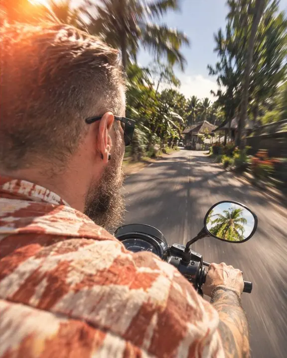 Seen from the passenger seat, a man rides a scooter with his back to the lens, the Bali road smeared ahead