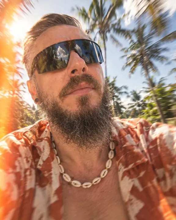 Close-up of a bearded man in sport shield sunglasses reflecting palm trees, an orange light leak at the edge