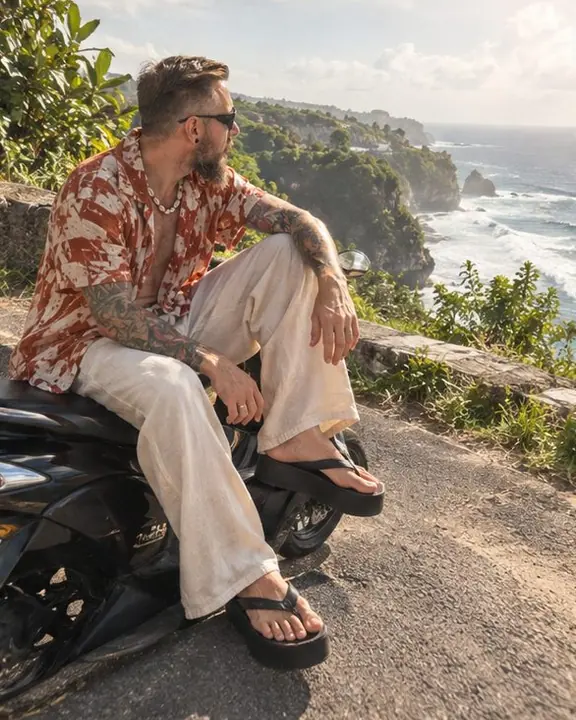 A bearded man sits on a parked scooter by the road, platform flip-flops on the asphalt, everything sharp