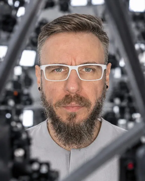 Through an outer dome lattice, a bearded man's face is framed by metal struts and small camera bodies