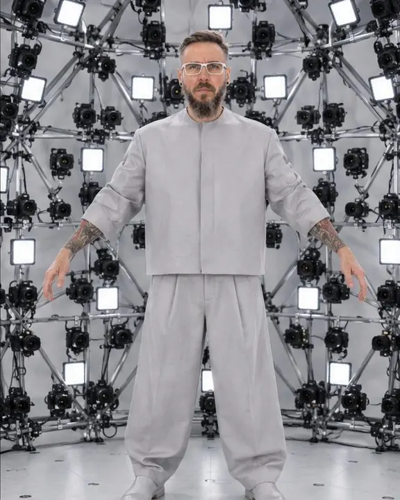 A man in silver-gray tailoring stands with arms slightly raised in even, shadowless light inside a scanning dome
