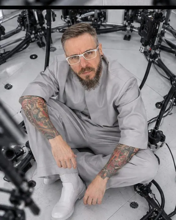 A bearded man sits on the dome floor among black cables, looking up at the camera-studded lattice