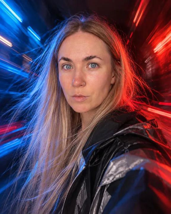 A woman with long blond hair, face sharp, blue light on one cheek and red on the other, light streaks radiating behind