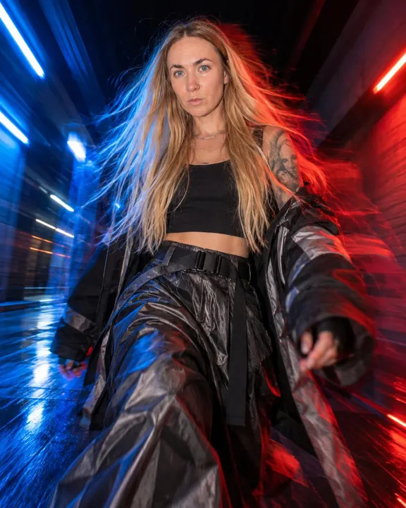 A woman with long blond hair walks at the lens in a black crop top and belted shiny trousers, red and blue trails behind