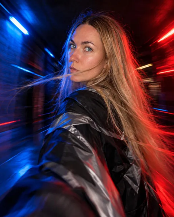 A woman with long blond hair looks back over her shoulder, a strand across her lips, blue light at left and red at right