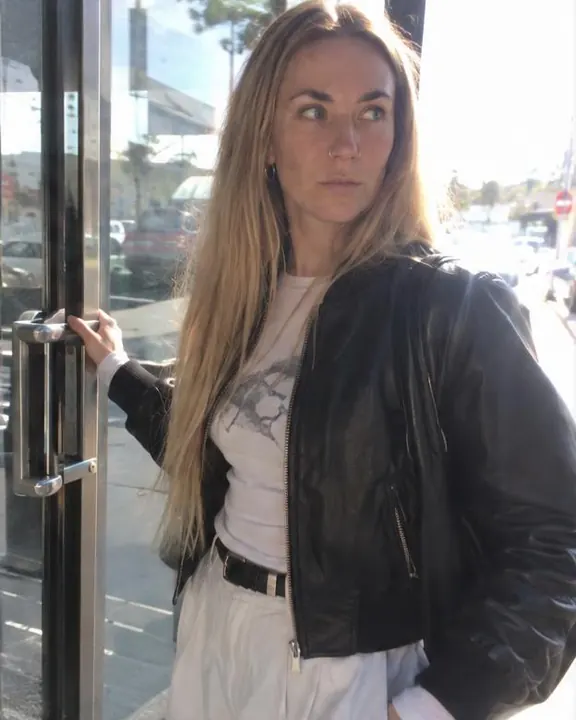 A woman with long blond hair pushes a glass door's steel bar mid-step, the bright street reflected as a white sheet