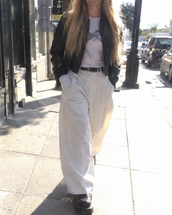 A woman with long blond hair walks toward the camera on a sidewalk, her cropped leather jacket shining hard in the sun