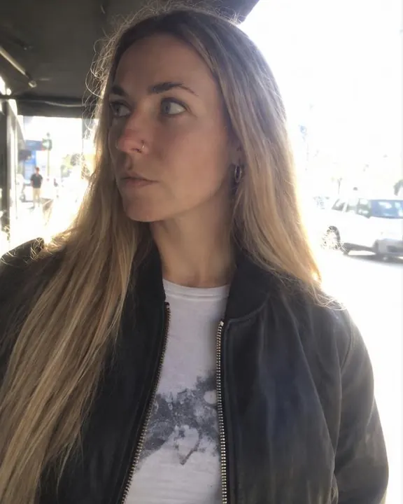 A woman with long blond hair stands under an awning looking aside, the sunlit street blown white with a cyan edge