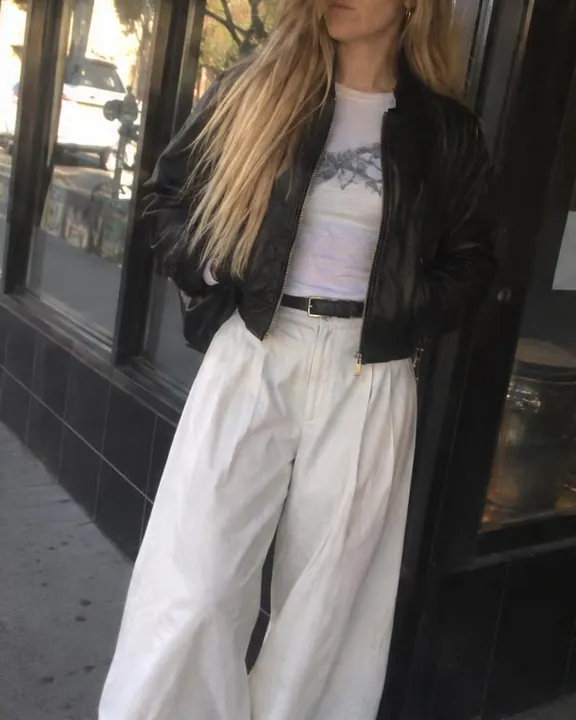 Crooked hip-height view of a woman with long blond hair by a shop window, hands in jacket pockets, feet cut at the edge