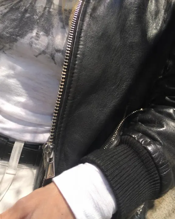 Phone close-up of a black leather jacket zip and cuff over a white sleeve, sun highlights clipped flat, smudgy shadows