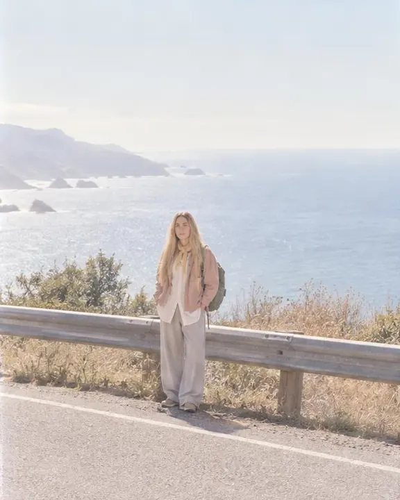 Wide view across an empty coast road: a woman with long blond hair stands small at the guardrail, sea and hills above