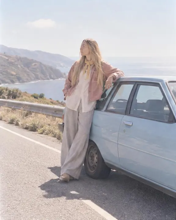 A woman with long blond hair in a dusty-pink jacket stands beside a pale-blue 1980s hatchback, one hand on its roof