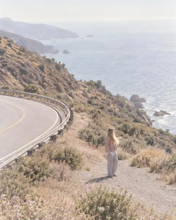 High view of a curving coast road, a woman with long blond hair small on the gravel shoulder, her ochre scarf lifting