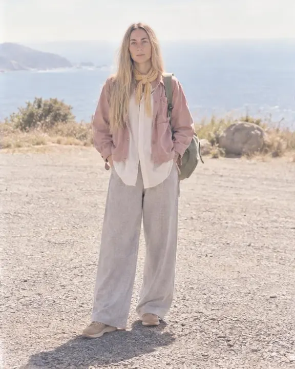 Full length of a woman with long blond hair standing square in a gravel lay-by, her long ecru shirt moving in the breeze
