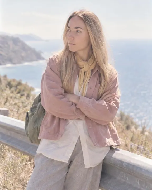 A woman with long blond hair leans back on a metal guardrail, arms folded, looking past the camera toward a pale sea