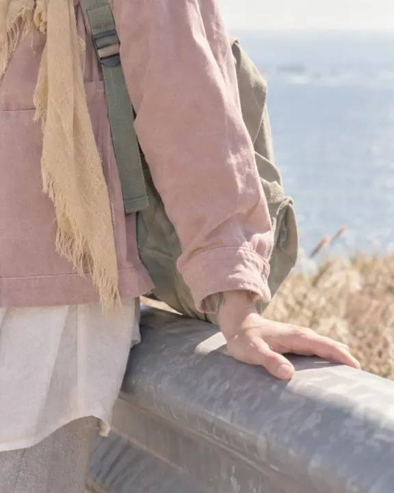 A hand on a sun-warmed metal guardrail beside a sage backpack strap and the frayed end of a cotton scarf, in soft pastels