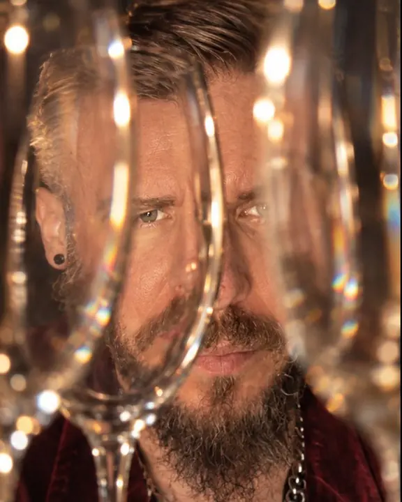 Through a row of wine glasses, a bearded man's face bends into curved vertical slices with one eye clear