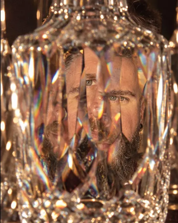 Through a cut-glass decanter, a man's face multiplies into faceted pieces edged with rainbow fringes