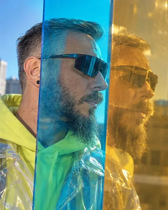 A bearded man's face reflected gold on a dichroic panel while also seen cyan through the same sheet