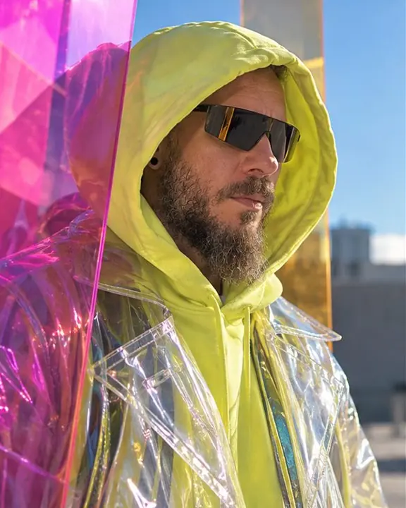 A man with an acid-yellow hood up, cyan and magenta light falling across his transparent PVC coat