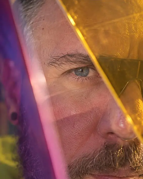 Close-up of one eye of a bearded man seen through the gold corner of a dichroic acrylic panel
