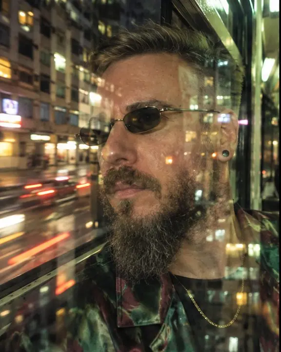 A bearded man's face against a tram window, building reflections over his cheek, the night street smeared outside