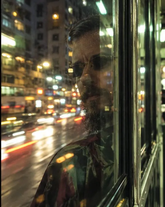 Reflection of a bearded man in a tram window layered over passing streaks of Hong Kong street lights