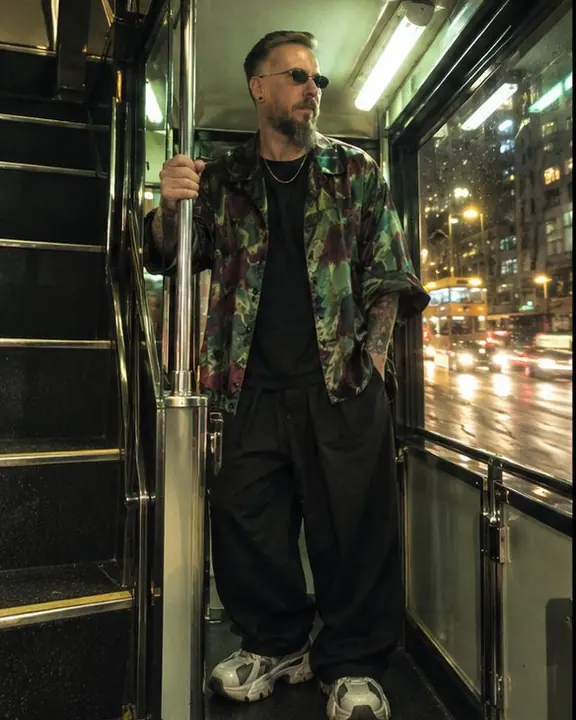 A man in a green and burgundy satin jacket stands by the tram stairs under a green-white fluorescent tube