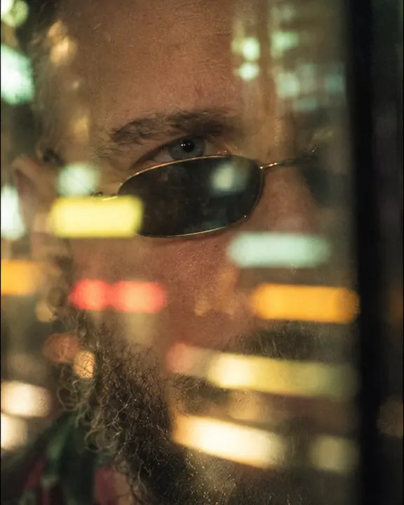 Close-up of one eye of a bearded man seen through blurred reflections of colored street lights on glass