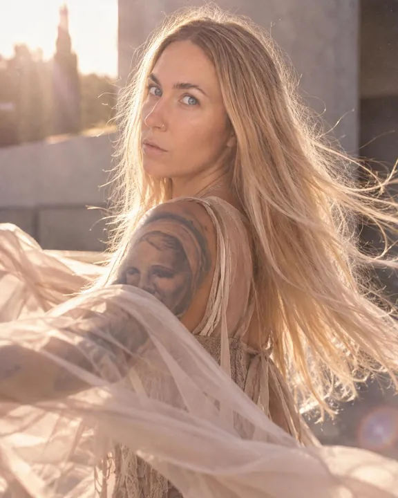 A woman with long blond hair turns over her shoulder, backlit hair flying, sheer fabric sweeping across the foreground