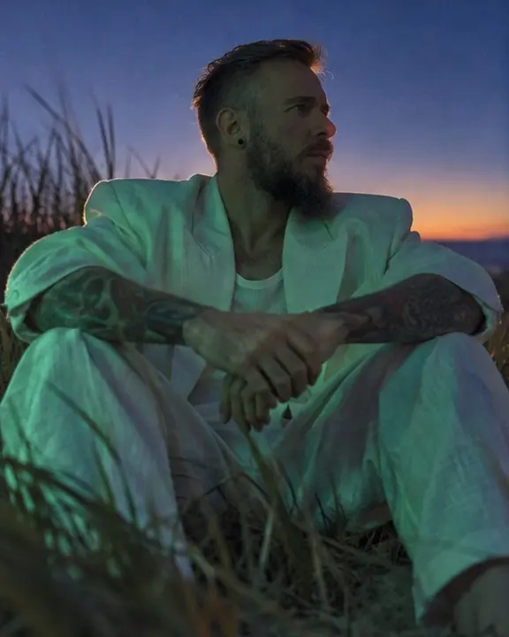 A man in a white suit sitting in a sand dune with knees up, green light falling across the linen