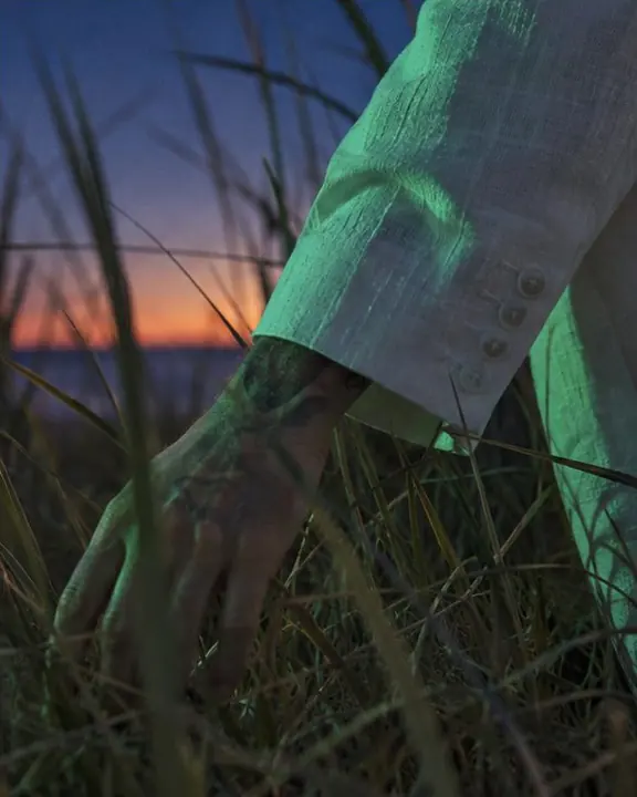 Detail of a white linen sleeve lit emerald green, surrounded by blurred dune grass at dusk