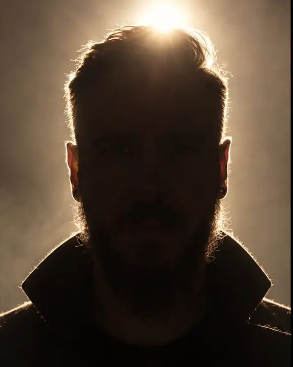 Frontal view of a man with light behind his head forming a corona, his face a dark disc with faint eyes