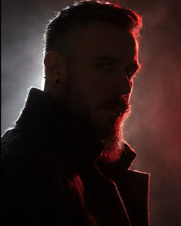 Three-quarter view of a bearded man in shadow, a cold white rim on one side and a deep red rim on the other