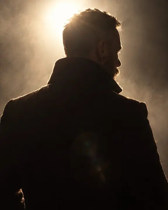 Back view of a man in a black coat looking over his shoulder into the light, his beard edge glowing
