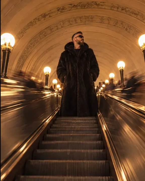 View from below of a long rising escalator, a figure in a floor-length fur coat at the top against an ornate vault