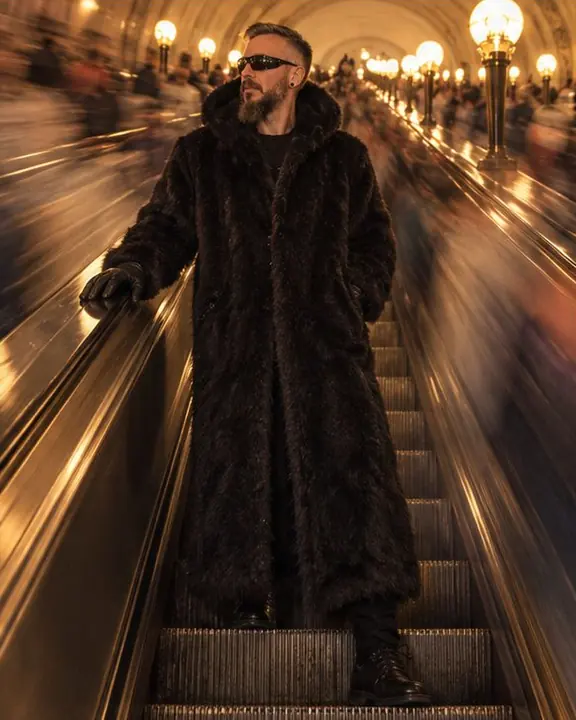 A man in a full-length black fur coat rests a gloved hand on the moving rail as the crowd streaks past
