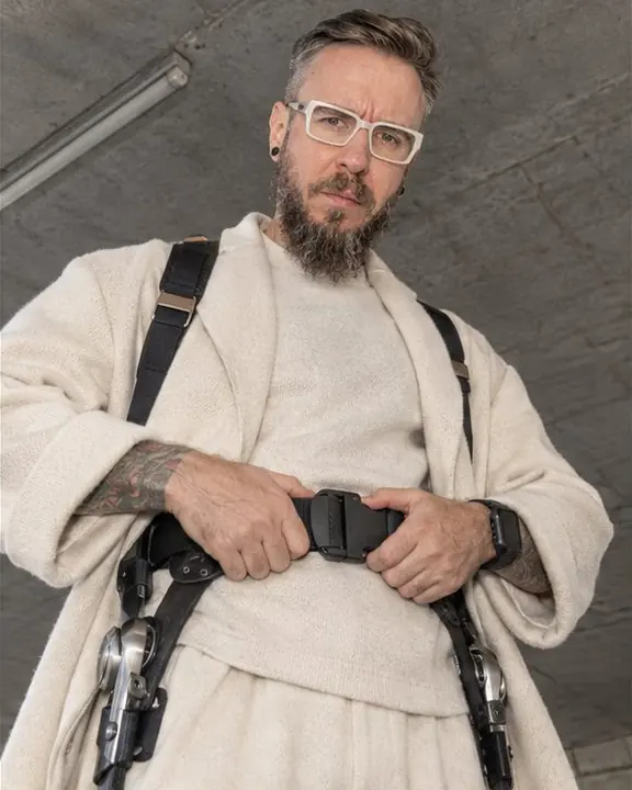 A bearded man in a cream cashmere coat adjusts the hip belt of a carbon exoskeleton, looking down at the camera