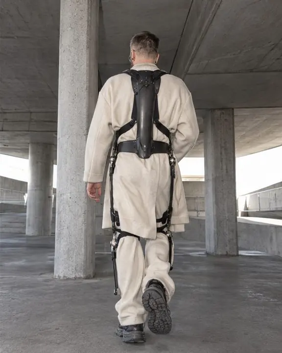 Back view of a man walking between concrete columns, a carbon spine plate strapped over his oversized cream coat