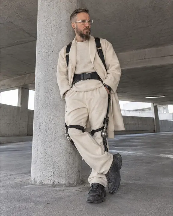 A man leans on a concrete column in a parking garage, wearing a slim carbon exoskeleton over cream cashmere