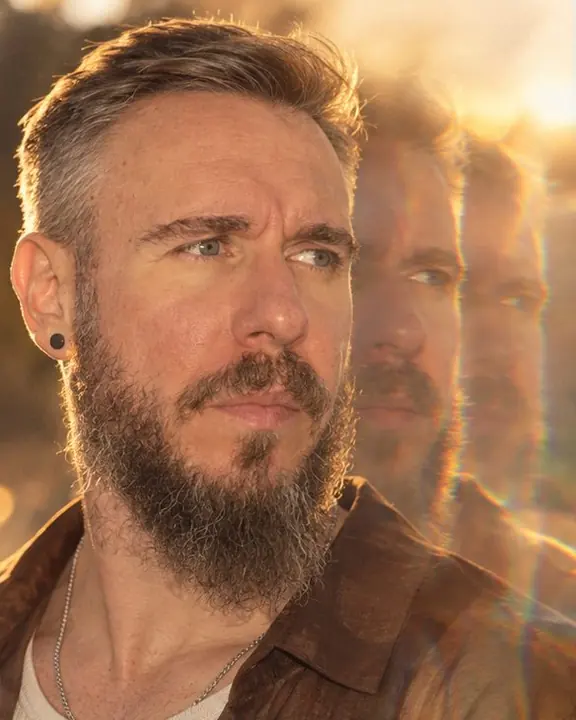 Tight portrait of a bearded man at golden hour, face three-quarters and sharp, two prism echoes drifting right