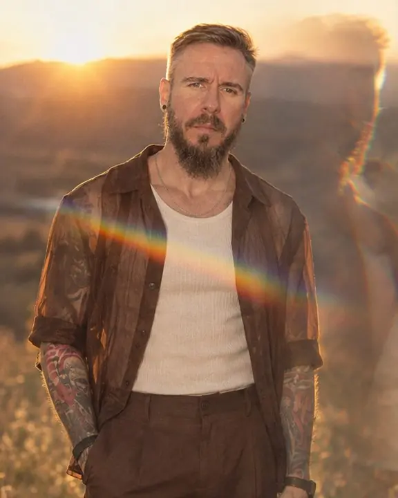 A man stands against the setting sun, a rainbow flare streak across his chest, a faint echo of his silhouette